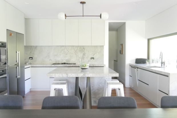 Bright contemporary open-plan kitchen and dining area with white full-height cabinetry, integrated stainless-steel appliances, a large marble-look island with waterfall edge, white stools, grey upholstered dining chairs, and a linear globe pendant. A matching marble-look splashback and benchtop continue to the sink run beside a large window. Warm timber flooring contrasts with the predominantly white and grey palette.