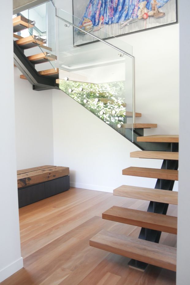 Bright contemporary stairway with floating timber treads supported by a dark steel stringer and enclosed by clear glass balustrades. The stair rises through a light-filled void, with warm timber flooring below and built-in dark cabinetry topped with a chunky timber bench. A colourful framed artwork is mounted high on the white wall, and greenery is visible through the stair opening.