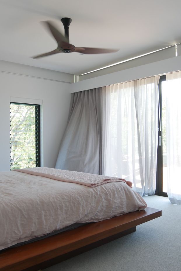 A bright, serene bedroom with a low timber platform bed dressed in pale textured bedding, white walls, soft grey curtains and sheer drapes. Large glazed doors and a louvred window bring in abundant natural light, while a sculptural ceiling fan provides a contemporary focal point.