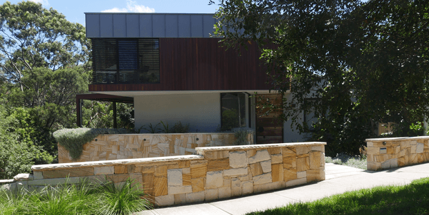 Exterior view of a contemporary multi-level house set among mature trees. The building combines dark standing-seam metal cladding, warm vertical timber wall cladding, extensive glazing with horizontal louvres, and a projecting upper storey over a partially open ground level. Layered rough-cut sandstone retaining and boundary walls frame a paved approach and landscaped foreground.
