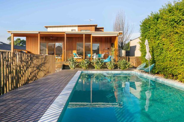 A contemporary timber-clad single-storey home with broad roof overhangs, a covered outdoor entertaining deck, and large glazed openings overlooking a swimming pool. The pool is bordered by timber decking, planted garden beds, and a vertical timber privacy fence.