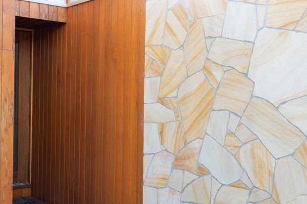 Close-up architectural detail showing a dark timber-lined doorway or entry recess beside an irregular natural stone wall with pale beige, cream, and warm tan tones. The composition focuses on contrasting vertical wood panelling and rough geometric stonework.