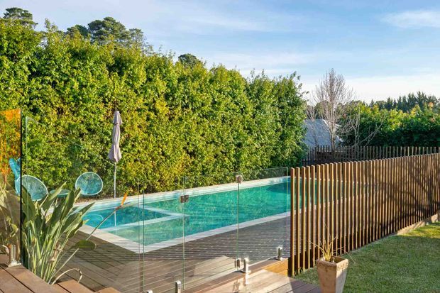 A landscaped residential pool area with a turquoise swimming pool enclosed by frameless glass balustrades, surrounded by a timber deck and vertical timber privacy screen. Dense greenery, outdoor furnishings, and a closed umbrella create a private garden setting.