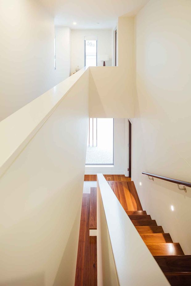 A bright, minimalist interior view of a modern stairway and landing. Warm timber stairs and flooring contrast with crisp white walls, angular balustrade walls, and a simple metal handrail. A tall glazed opening brings natural light into the multi-level circulation space.
