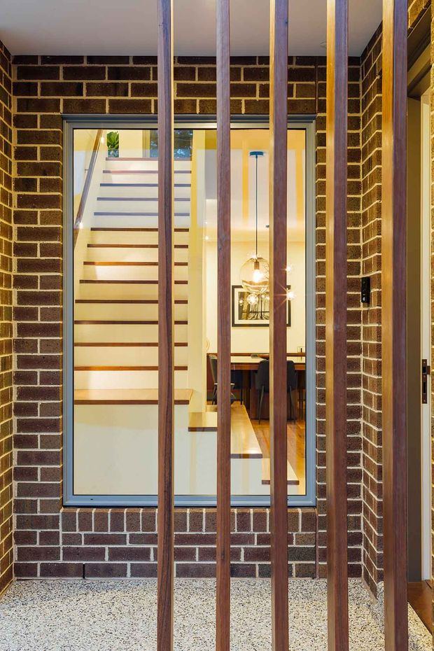 View from an entry or transitional area through vertical timber screening toward a glazed interior. Dark exposed brick walls frame a large window, with a contemporary timber staircase, warm timber flooring, dining furniture, and pendant lighting visible beyond. The foreground has a speckled terrazzo-like floor and the composition highlights layered thresholds, brickwork, glazing, and timber slats.