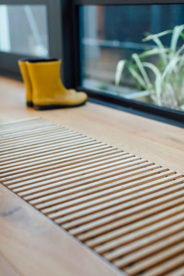 Close-up interior detail of a light timber slatted floor mat or grate beside a large dark-framed window. A bright yellow rubber boot rests near the glazing, with leafy planting visible outside. The image emphasises natural timber tones, linear detailing, and a practical entry or threshold area.