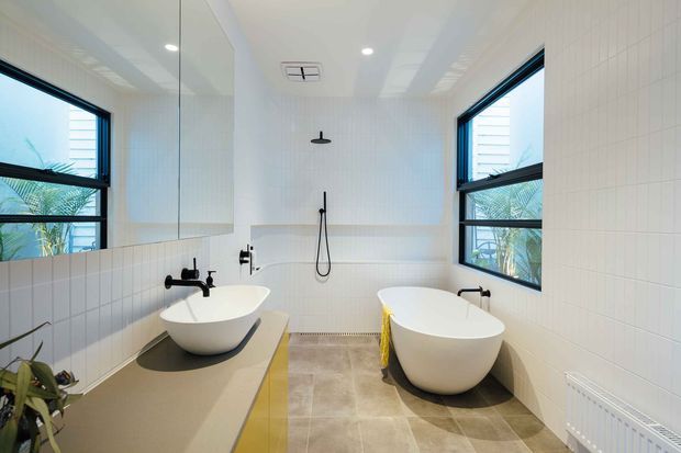 Bright contemporary bathroom with twin freestanding oval basins on a long vanity, two freestanding bathtubs, wall-mounted black fixtures, white tiled walls, large black-framed windows, and a full-width mirror. The room has a clean, symmetrical layout with warm neutral floor tiles and abundant natural light.