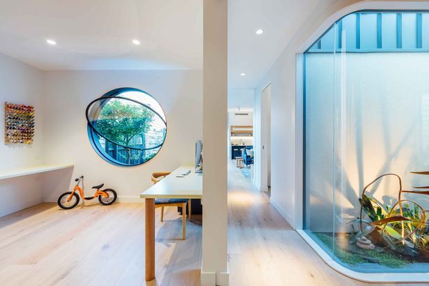 Bright contemporary interior showing a compact workspace or study area opening onto a hallway. Pale timber flooring, white walls, recessed ceiling lights, a simple desk with timber legs, bicycles, and a large circular window provide the main visual features. The hallway includes a dramatic curved, floor-to-ceiling glazed internal feature with plants and blue-tinted glazing, while a distant blue seating area is visible beyond.