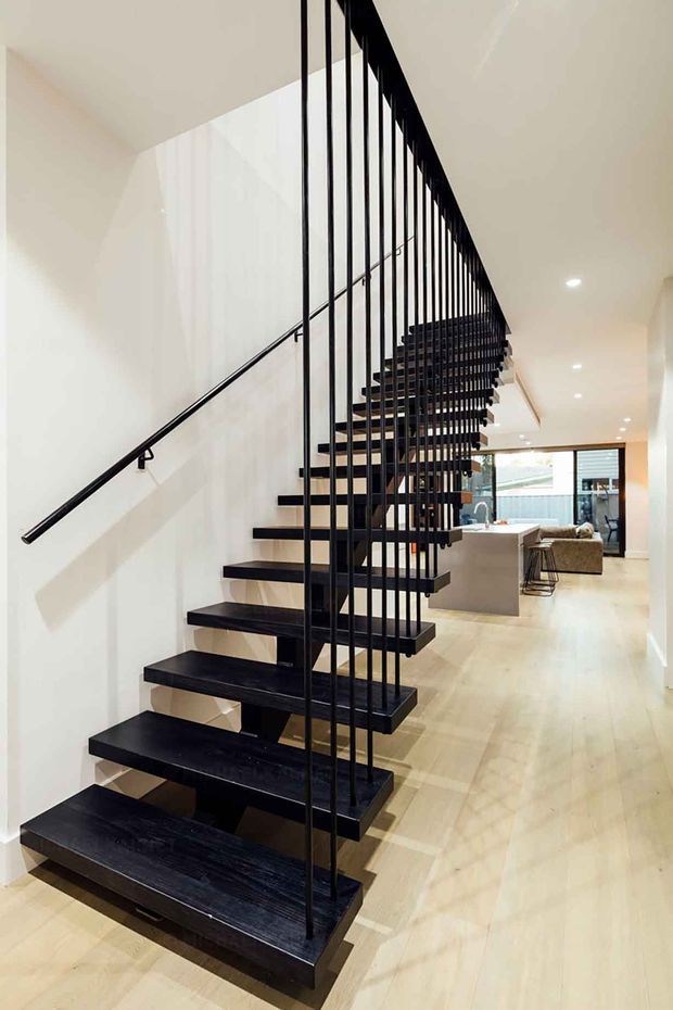 Interior view of a contemporary open-riser staircase with dark timber or painted-black treads and slim black metal vertical balusters. The stair runs beside a white wall and is integrated into a bright open-plan home, with pale timber flooring, recessed ceiling lights, and a kitchen/living area visible beyond.
