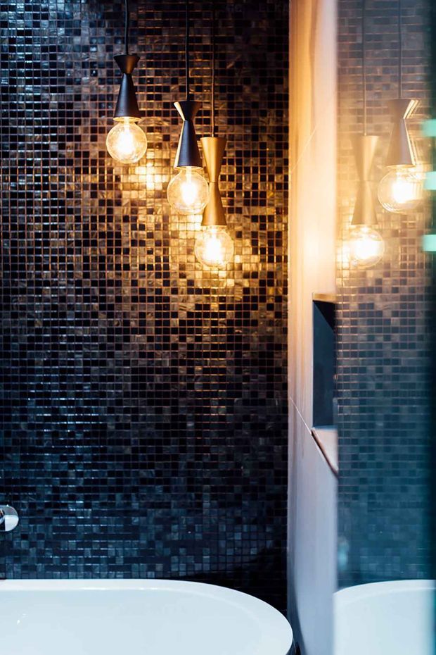 A dramatic bathroom interior featuring a freestanding white bathtub, a full-height wall clad in small glossy dark mosaic tiles, and three sculptural pendant lights with glowing exposed bulbs. A large mirror on the right reflects the tiled wall and lighting, adding depth. The palette is predominantly deep blue-black, bronze, warm gold, and white.