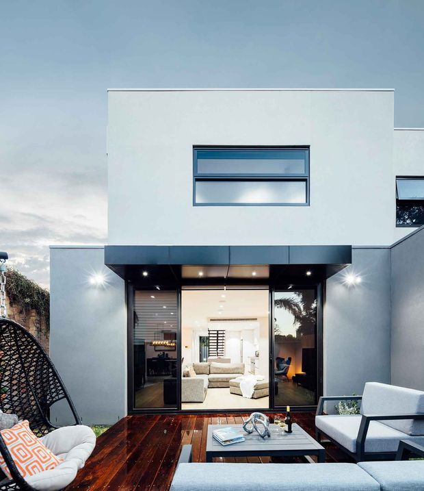A contemporary two-storey residential facade viewed from an outdoor timber deck. The smooth white rendered building has a boxy flat-roof form, dark-framed horizontal windows, a deep dark canopy, and large glazed sliding doors opening to a bright living room. The deck is furnished as an outdoor lounge with sofas, a coffee table, and a hanging chair, with warm evening lighting and a palm visible through the glazing.