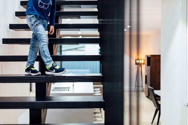 Interior view of a contemporary open-riser staircase with dark timber or metal treads and a substantial dark central support, leading through a bright multi-level space. A child is walking on the stairs; a minimalist floor lamp, dark furniture, and a timber cabinet are visible in the adjacent area.