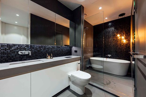Contemporary bathroom with a floating white vanity, integrated basins, wall-mounted tapware, and a wall-hung toilet. Mirrored cabinetry and glass shower screening reflect the room, while a freestanding white bathtub sits against a dramatic black mosaic feature wall illuminated by warm pendant lighting. Grey floor tiles and dark accents create a polished, high-contrast finish.