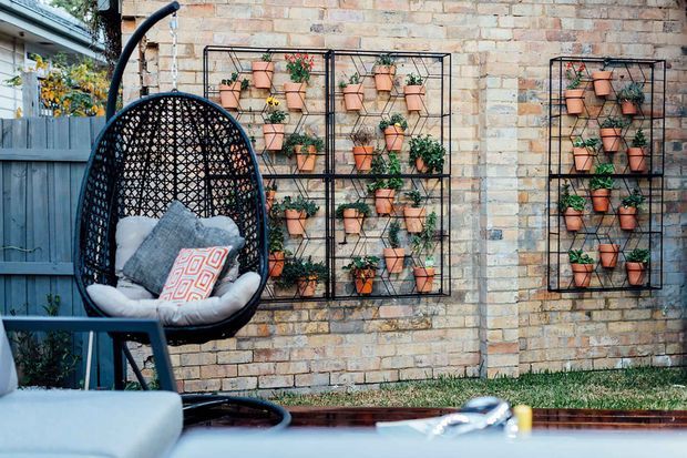 A compact outdoor courtyard or patio seating area with a hanging egg chair in the foreground, positioned beside a warm exposed-brick wall. Two black metal vertical plant racks hold rows of terracotta pots and greenery, creating a garden feature wall. A dark privacy fence and neighbouring structure are visible to the side, with a low pool or water feature edge along the bottom of the image.
