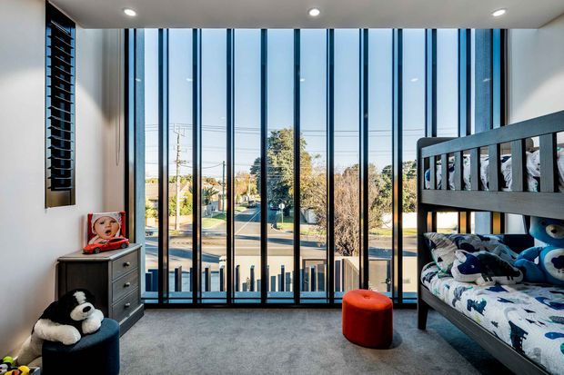 Bright, contemporary children's bedroom with bunk beds, grey carpet, a dark chest of drawers, plush toys, and a red ottoman. Floor-to-ceiling vertical glazing fills the rear wall, providing abundant daylight and views to the street, while a high horizontal window adds ventilation and light on the side wall.