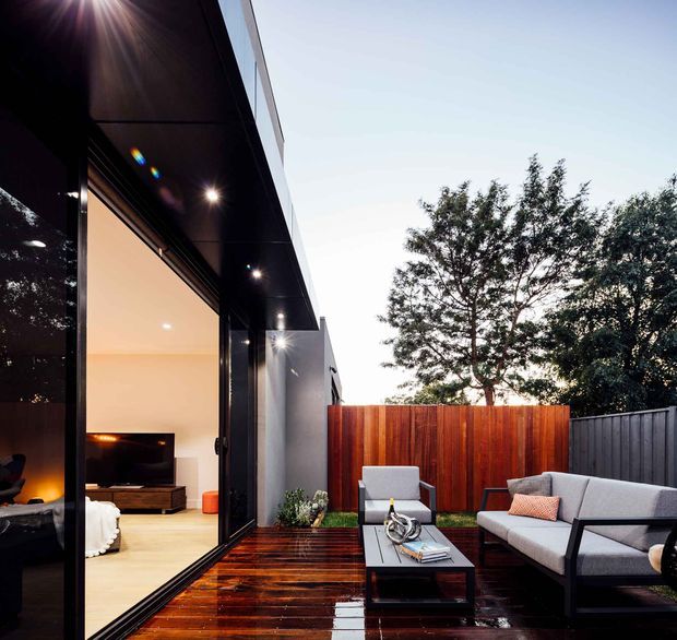 Contemporary single-level home opening onto a compact outdoor entertaining terrace. Large black-framed glazed doors connect the warmly lit living room to a dark timber deck furnished with a sofa, armchairs and low coffee table. A vertical timber privacy fence and dark boundary fence enclose the space, with mature trees visible beyond. The image is photographed from the exterior and primarily focuses on the furnished deck and indoor-outdoor connection.