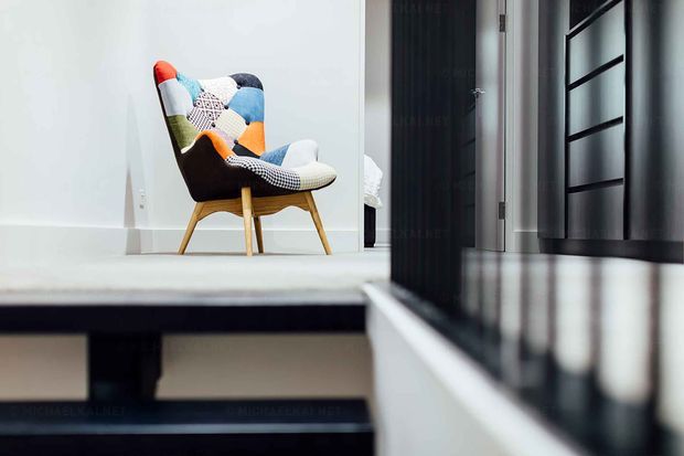 A minimalist interior view of a small landing or hallway beside a short set of dark-edged steps. A colourful patchwork lounge chair with timber legs sits against a pale wall, while a black-framed glazed or reflective partition and dark doorway line the right side. The scene has a clean, contemporary feel with strong horizontal and vertical lines.