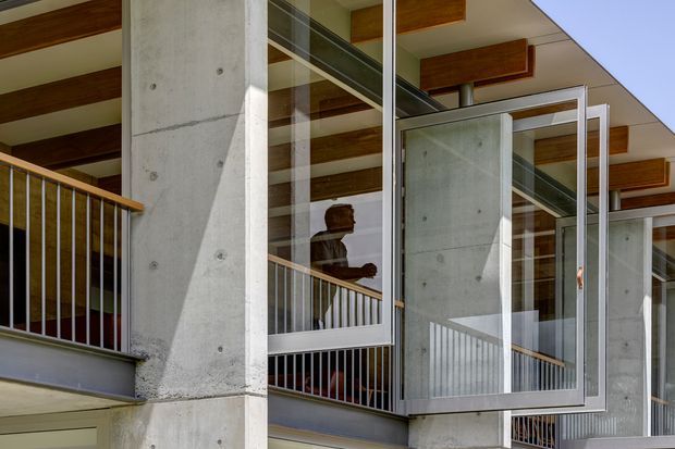 Exterior architectural detail of a contemporary multi-level building, featuring exposed concrete structural columns and walls, extensive glazed openings, metal balustrades, and timber-lined ceiling beams. A person is visible through the glazing on an upper-level walkway or balcony.