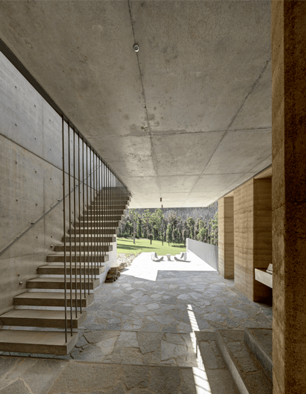 A covered open-air circulation space with a raw concrete ceiling and wall, a floating stair with slender metal balustrade, and irregular natural stone paving. Timber-clad or stone-clad built-in volumes line the right side, while the space opens toward a sunlit landscaped courtyard. The architecture has a strongly monolithic, minimalist character with exposed structural concrete and carefully framed outdoor views.