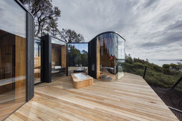 Contemporary pavilion-like coastal retreat with curved floor-to-ceiling glazing, dark vertical cladding, warm timber interiors and a broad timber deck overlooking a wooded landscape and distant water. The image highlights sculptural curved forms, reflective glass, and an outdoor soaking tub integrated into the deck.