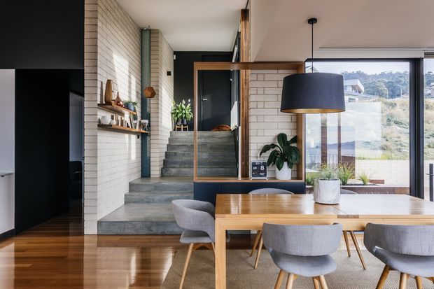 Contemporary open-plan dining and living space with a timber dining table and upholstered chairs, polished timber flooring, exposed pale brick walls, and a large black pendant light. Concrete steps lead to a raised hallway or living area, while expansive glazing provides views of the surrounding landscape.