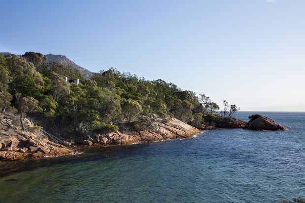 A scenic coastal landscape showing a rocky, tree-covered shoreline extending into calm blue water, with rugged hills and distant open sea under a clear sky. No building or architectural structure is visible.