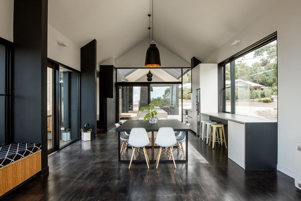 Contemporary open-plan dining and kitchen interior with a vaulted ceiling, dark polished concrete flooring, extensive black-framed glazing, and a minimalist dining table with white chairs. Black pendant lighting, dark cabinetry, and a compact kitchen with bar stools create a refined monochromatic scheme, while trees and neighboring buildings are visible outside.