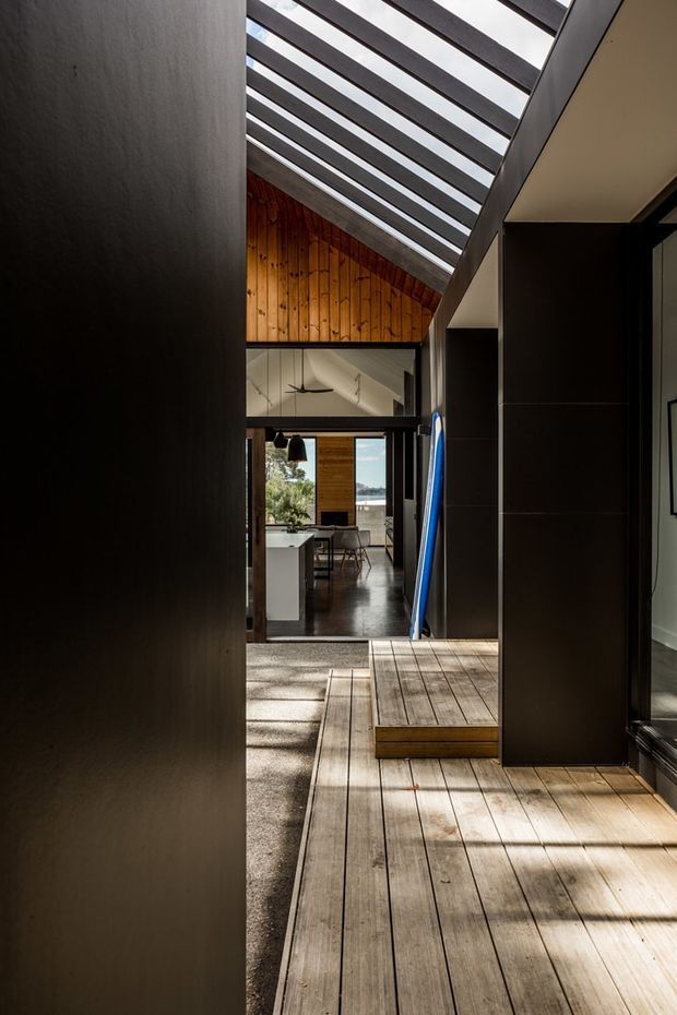 A narrow, light-filled transitional passage inside a contemporary home, with warm timber decking, dark built-in wall panels, and a dramatic slatted roof or clerestory overhead. The view leads toward an open-plan kitchen and dining area, with timber wall cladding and glazing beyond. Strong linear shadows and exposed structural framing create a high-contrast architectural composition.