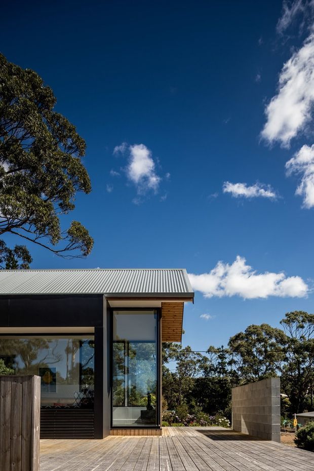 Exterior architectural photograph of a compact contemporary house set among trees. The visible structure has a simple rectangular form, dark cladding, extensive floor-to-ceiling glazing, a light corrugated metal roof, timber soffit, and a broad timber deck enclosed partly by concrete block walls. Bright blue sky and surrounding vegetation frame the building.