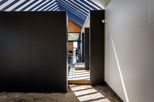 A narrow, dramatic interior passage with tall matte-black built-in partitions and cabinetry, a white paneled wall, and a striking blue-and-white striped vaulted ceiling. Strong sunlight creates geometric shadows across the pale textured floor, while a timber-lined area and glazed opening are visible at the far end.