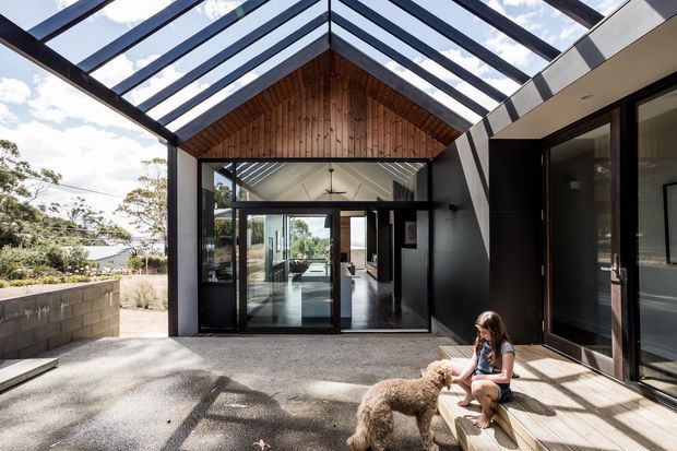 A contemporary residential courtyard or terrace framed by a dramatic glazed gable roof. Dark metal-framed glazing and cladding contrast with a warm vertical timber-lined gable, while the covered outdoor area connects directly to the bright interior living spaces. A low masonry wall and restrained landscaping sit to the left, with timber decking and steps to the right.