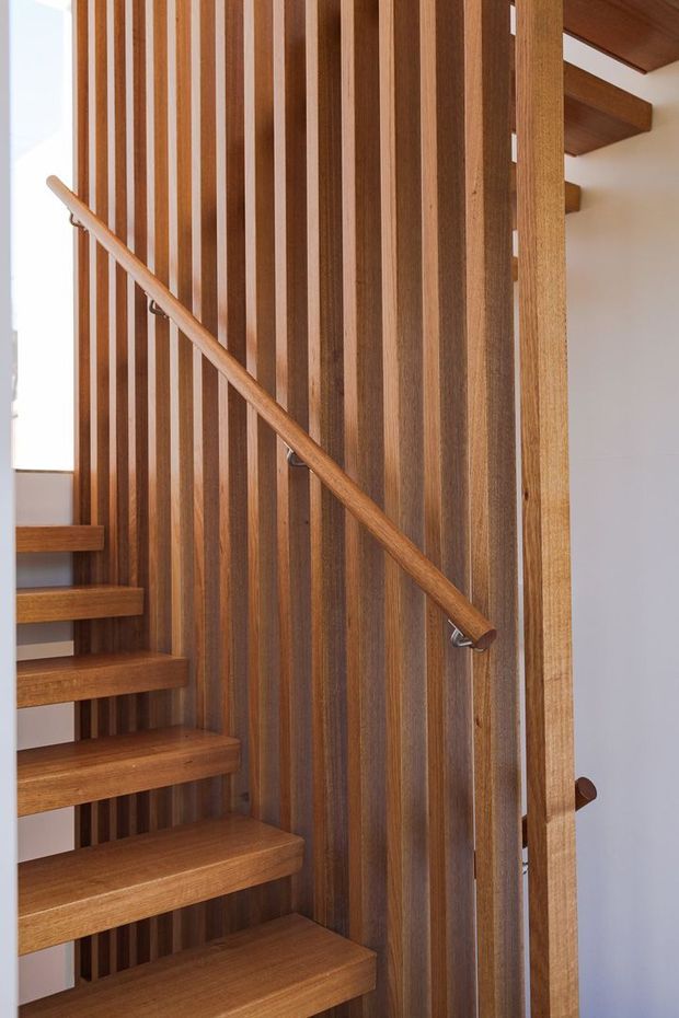 Close-up interior view of a contemporary timber staircase with open-riser wooden treads, a simple timber handrail, and a vertical timber slat screen forming the stair balustrade and feature wall. White plaster walls and strong natural light create a warm, refined stairwell.