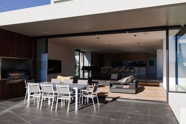 Modern open-plan indoor-outdoor entertaining area with a large tiled terrace, outdoor dining setting, built-in barbecue, and wide retractable glazing opening into a contemporary living room and kitchen. The interior features timber flooring, dark timber wall joinery, a neutral sectional sofa, and a kitchen island. Strong indoor-outdoor connection and clean-lined architectural detailing are evident.