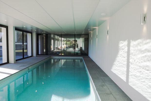 A bright, modern indoor swimming pool corridor with a long turquoise pool, pale tiled surround, white walls and ceiling, and extensive black-framed glazing at one end and along the side. Strong daylight creates reflections on the water and wall, with a minimalist, clean architectural character.