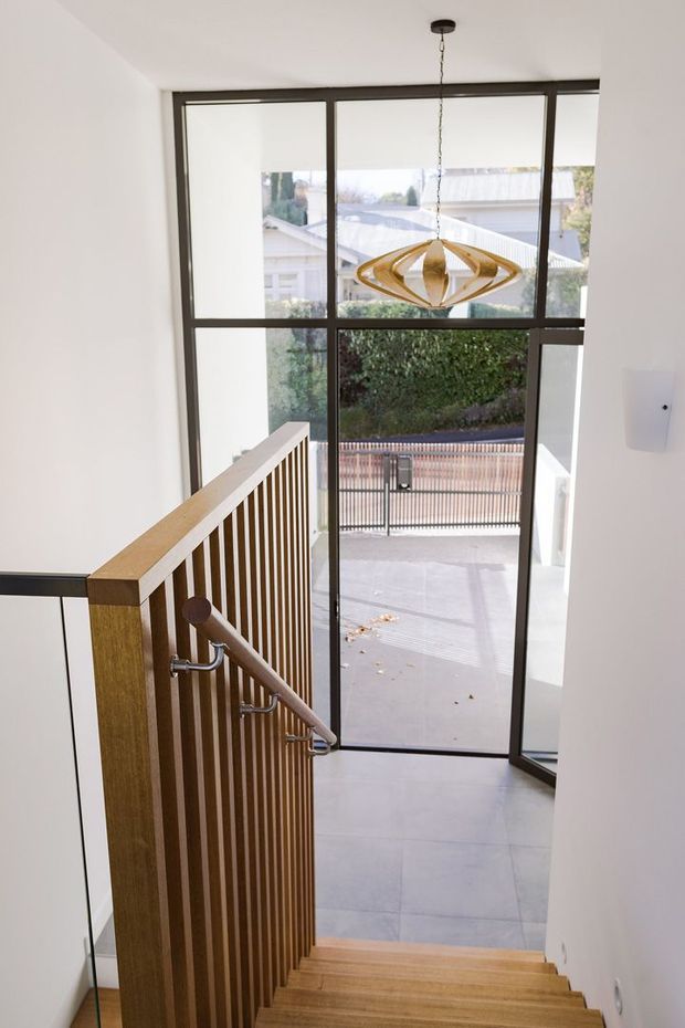 Bright contemporary stairway descending toward a glazed entry door. The stair has warm timber treads and a timber handrail with vertical timber slats and clear glass balustrade panels, paired with dark metal window and door frames. A sculptural pendant light hangs in front of the full-height glazing, with an exterior driveway and neighbouring buildings visible beyond.