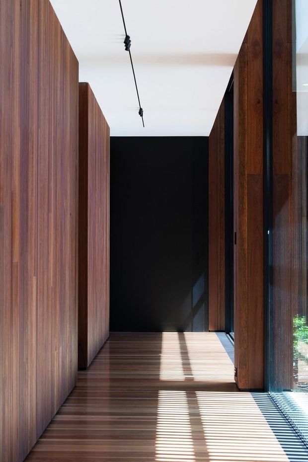 A narrow, refined interior corridor lined with floor-to-ceiling dark timber wall panelling and matching timber flooring. A black feature wall anchors the far end, while full-height glazing on the right brings in strong sunlight and casts striped shadows across the floor. Minimal black track lighting runs along the white ceiling.