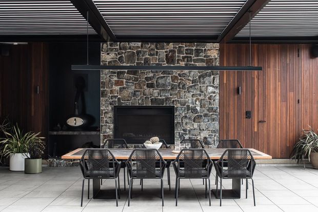 A sophisticated covered outdoor dining area with a long timber dining table and black woven chairs, arranged in front of a dark stone feature wall with a built-in fireplace. Vertical timber cladding, integrated cabinetry or doors, potted plants, and a slatted roof structure create a warm, refined contemporary entertaining space.
