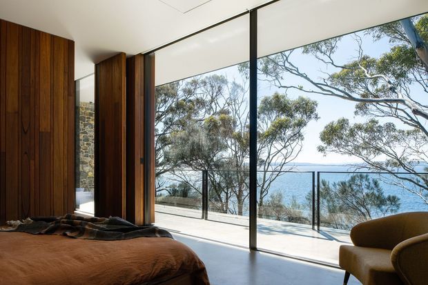A contemporary bedroom with expansive floor-to-ceiling glazing opening onto a balcony or terrace overlooking the ocean. Rich vertical timber wall panelling and timber-framed doors contrast with a pale smooth floor, while a low bed and curved upholstered armchair create a warm, refined interior. Dark slender glazing frames and a glass balustrade preserve the coastal view.