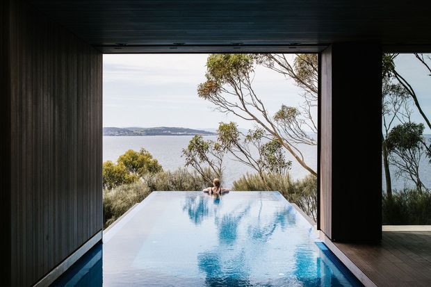 A dark, timber-lined pavilion opens directly onto a reflective infinity pool overlooking a calm body of water and a tree-covered shoreline. The strong indoor–outdoor connection, expansive opening, and natural setting create a secluded, dramatic retreat.