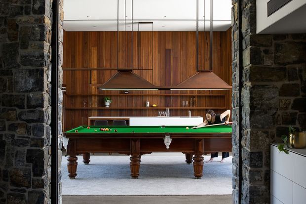 Interior view through a rough dark-stone opening into a sophisticated games room featuring a full-size green-felt billiards table, rich vertical timber wall panelling, built-in shelving, two large copper-toned pendant lights, and a grey carpeted floor. A partial white kitchen or utility joinery area is visible at the right edge, while people are present in the background.