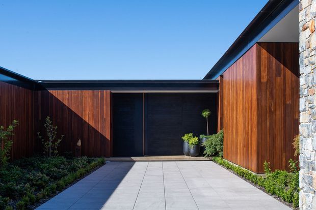 A modern residential courtyard or entry approach framed by dark vertical timber-clad walls and a black feature garage or entry door. Large pale rectangular paving slabs lead through low landscaped planting, with a contrasting pale rough-stone wall and a sharply angled roofline visible on the right. Strong sunlight creates graphic shadows across the timber facade.