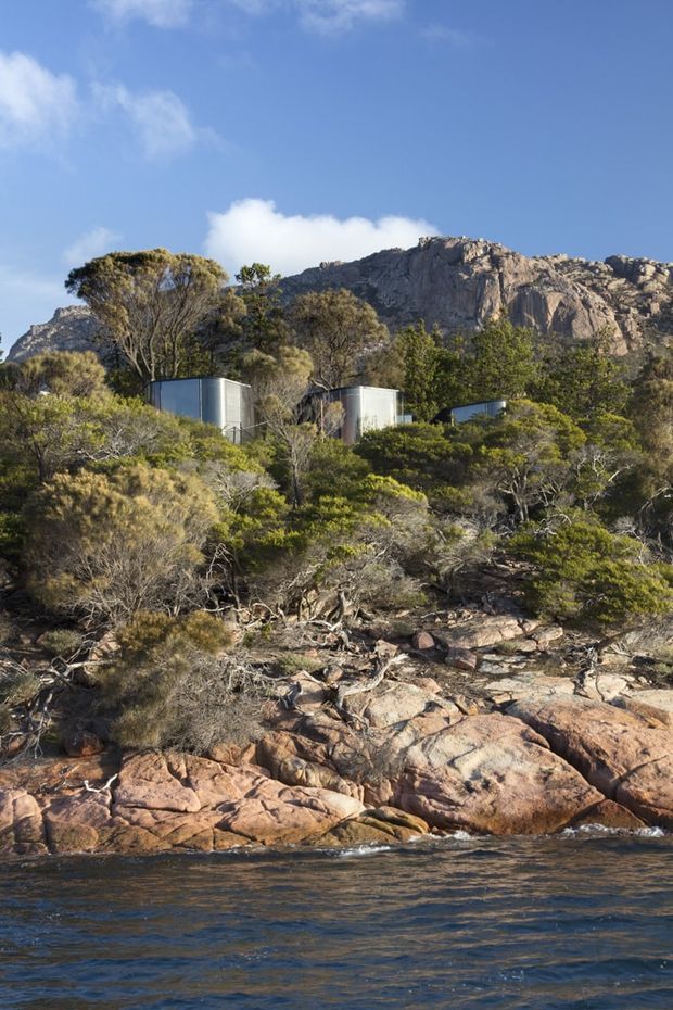A rugged coastal or lakeside landscape with distinctive rounded rock formations, native scrub vegetation, and calm water in the foreground. Several small cylindrical or curved-roof metallic buildings are partially concealed among the trees, with a dramatic rocky mountain ridge rising behind them.