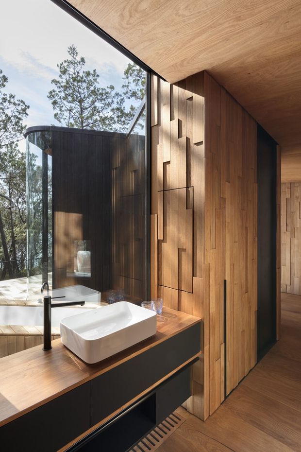 Contemporary bathroom with a sculptural white vessel basin on a dark built-in vanity, warm timber benchtop, extensive timber wall panelling and matching timber floor and ceiling. A freestanding white bathtub is visible beside full-height glazing, which reflects the surrounding trees and landscape. The room features black tapware, integrated dark cabinetry and a textured geometric timber feature wall.