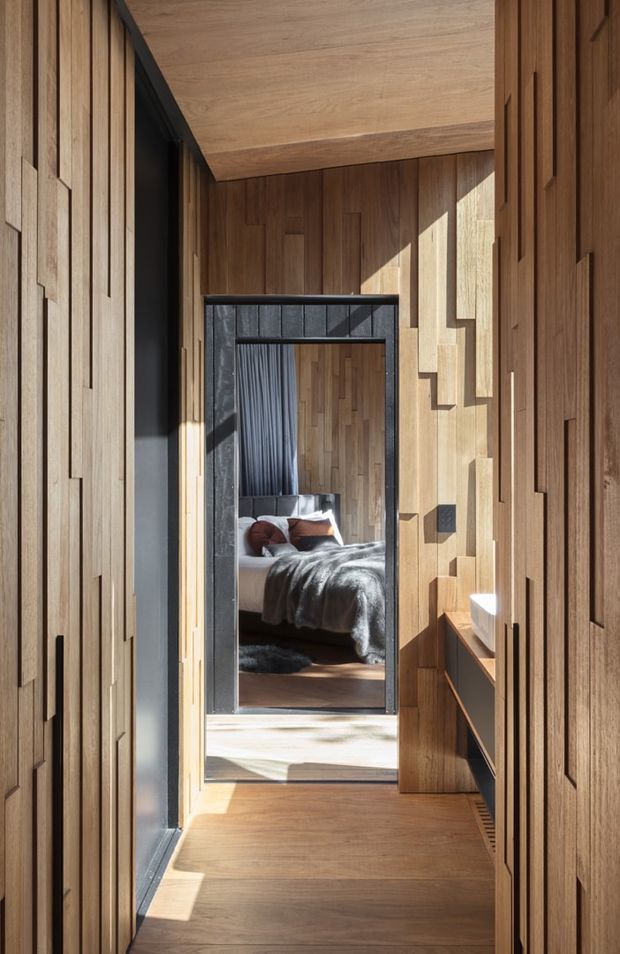 A narrow, richly timber-lined hallway with integrated vertical panelled joinery and warm timber flooring. A dark-framed doorway opens directly toward a bedroom visible beyond, where a bed, pillows, draped curtains and a dark throw can be seen. Strong sunlight creates geometric shadows across the timber walls and floor, while a slim built-in console runs along the right side.