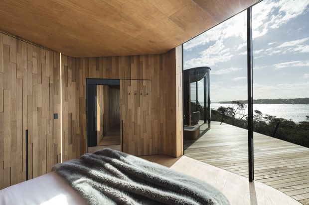 A contemporary bedroom clad in warm vertical timber wall panelling and a matching timber ceiling. The room opens through a dark-framed doorway toward a glazed outdoor area, with floor-to-ceiling glazing revealing a timber deck and a calm waterfront landscape. A bed with a dark textured throw occupies the foreground.