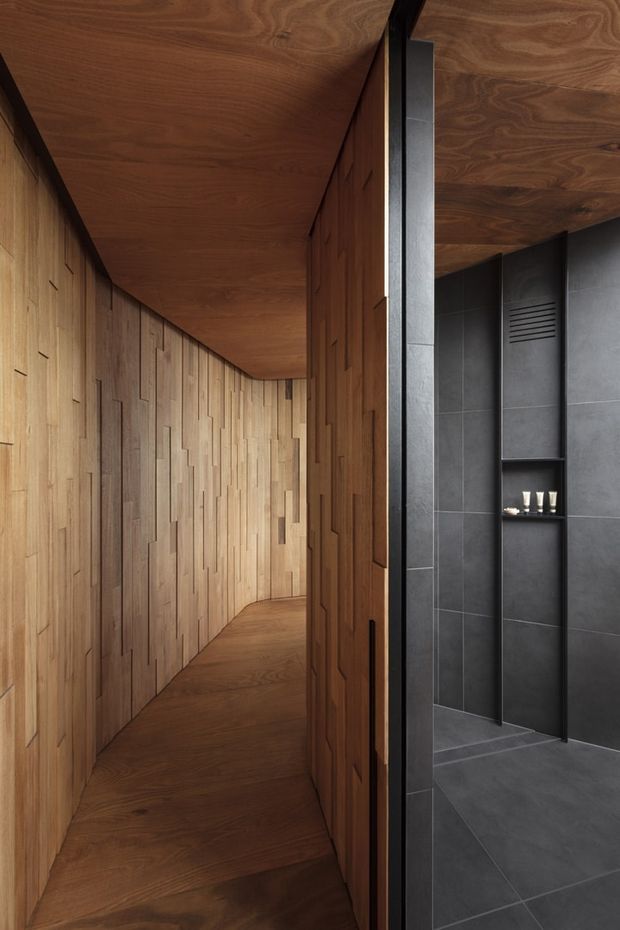 A narrow, refined interior passage clad extensively in warm timber, with sculptural vertical wall detailing and a matching timber ceiling and floor. At right, a partially visible dark charcoal tiled bathroom or shower area includes a recessed shelf and black fixtures. The design is minimalist and contemporary, with strong material contrast between the timber corridor and dark stone-like tiles.