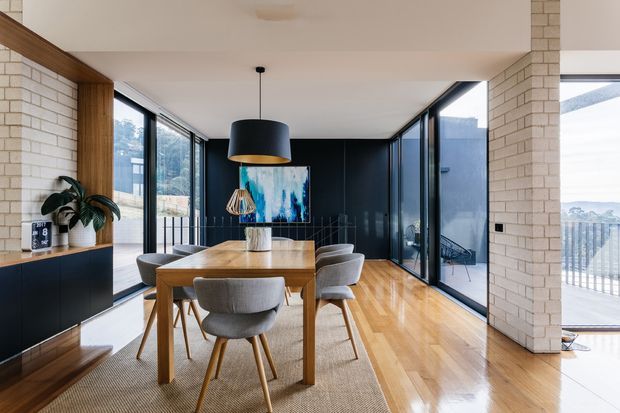 A bright, contemporary open-plan dining area with a timber dining table and upholstered chairs, framed by full-height glazing. White brick walls, warm timber flooring, dark built-in cabinetry, and a black feature wall create a refined contrast, with views to an exterior balcony or terrace.