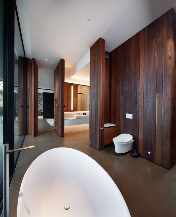 A spacious contemporary bathroom featuring a large freestanding white bathtub in the foreground, a wall-mounted white toilet, double vanity, warm integrated lighting, dark polished concrete flooring, and extensive vertical dark timber wall panelling and doors. Full-height glazing is visible at the left, contributing natural light and a connection to the outdoors.