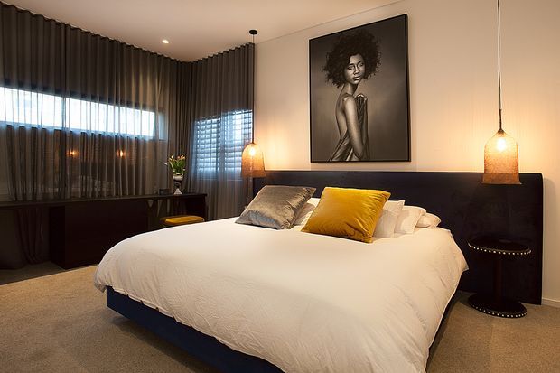 A sophisticated contemporary bedroom featuring a large upholstered bed with white bedding, dark navy or charcoal headboard and base, layered grey, white and mustard cushions, dark full-height curtains, a dark built-in sideboard, and warm woven pendant lights. A monochrome portrait provides a strong feature above the bed. The room has carpeted flooring, soft neutral walls, and a warm, moody atmosphere.
