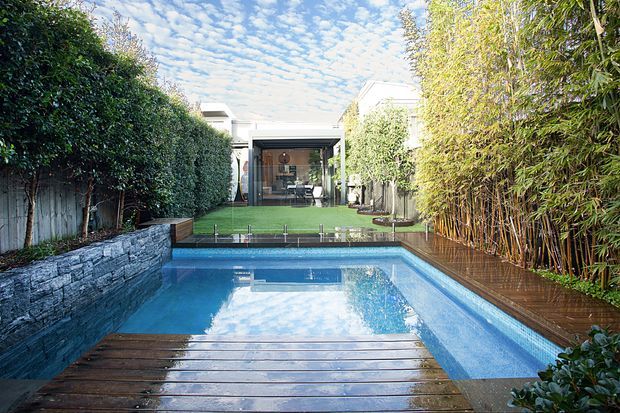 A landscaped residential courtyard viewed toward a rectangular swimming pool and timber deck. Dense clipped hedges and bamboo screening create privacy along both sides, with a low stacked-stone retaining wall bordering the pool. Beyond the lawn, a modern glazed pavilion or living area is visible. The scene is bright and tranquil, with strong reflections in the still blue water.